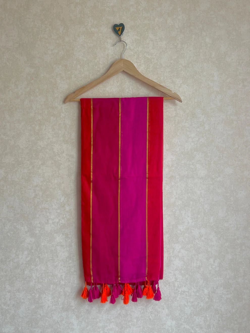Pink scarf with gold stripes and orange tassels on a wooden hanger against a beige wall.